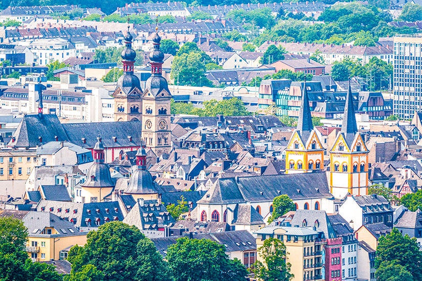 Eine dichte Stadtlandschaft mit vielen historischen Gebäuden und mehreren markanten Kirchtürmen prägt den Blick. Besonders auffällig sind Kuppeln und schlanke Spitzen, die aus den grauen und bunten Dächern herausragen. Im Hintergrund ziehen sich moderne Wohn- und Bürogebäude bis zum Horizont. Koblenz Stadt