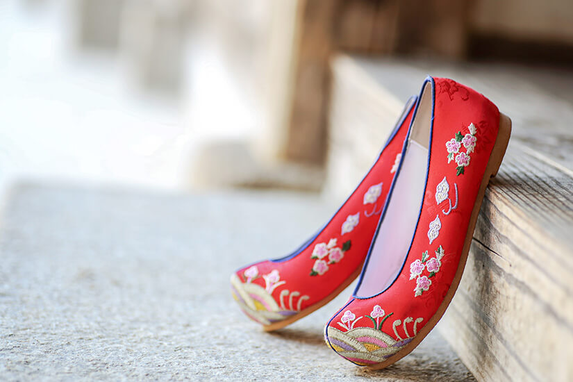 Rote, flache Schuhe lehnen an einer Holzstufe. Sie sind mit feinen Blumenmustern bestickt. Die Farben wirken kräftig und traditionell, mit dekorativen Details an der Spitze. Souvenirs Korea Kkotsin