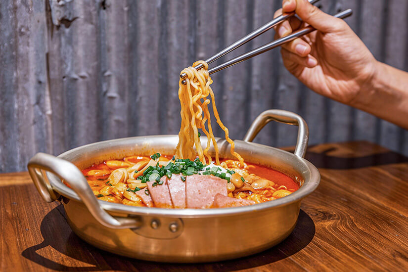 In einem Metalltopf köcheln Nudeln in einer roten, würzigen Brühe mit Toppings wie Frühlingszwiebeln und Wurstscheiben. Souvenirs Korea Ramyeon