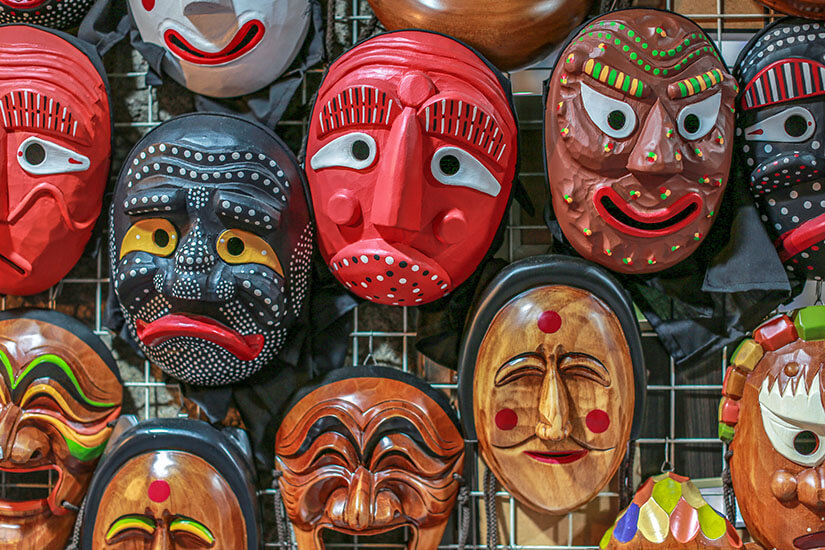 Mehrere bunte, geschnitzte Masken hängen dicht nebeneinander an einem Gitter. Die Gesichter haben stark überzeichnete Ausdrücke mit großen Augen, Mustern und kräftigen Farben. Souvenirs Korea Hahoe