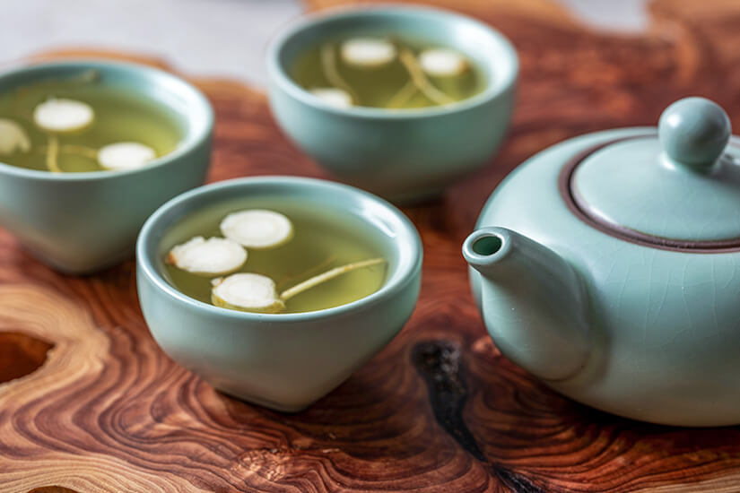 Mehrere kleine Keramikschalen mit hellem Tee stehen auf einer gemaserten Holzoberfläche. Darin schwimmen feine Scheiben Ginseng. Souvenirs Korea Tee