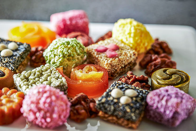 Auf einem Teller liegt eine bunte Auswahl kleiner, kunstvoll geformter Süßigkeiten und Snacks. Die Stücke haben unterschiedliche Texturen, von knusprig und körnig bis weich und fruchtig. Souvenirs Korea Hangwa