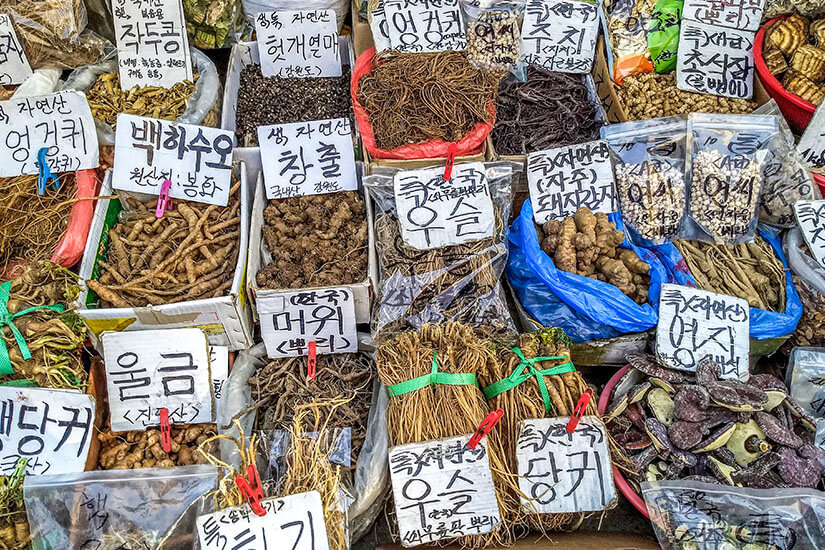 Viele Säcke und Boxen sind mit getrockneten Wurzeln, Kräutern und anderen Naturprodukten gefüllt. Handgeschriebene Schilder mit koreanischen Schriftzeichen kennzeichnen die einzelnen Waren. Natuerliche Medizin Korea
