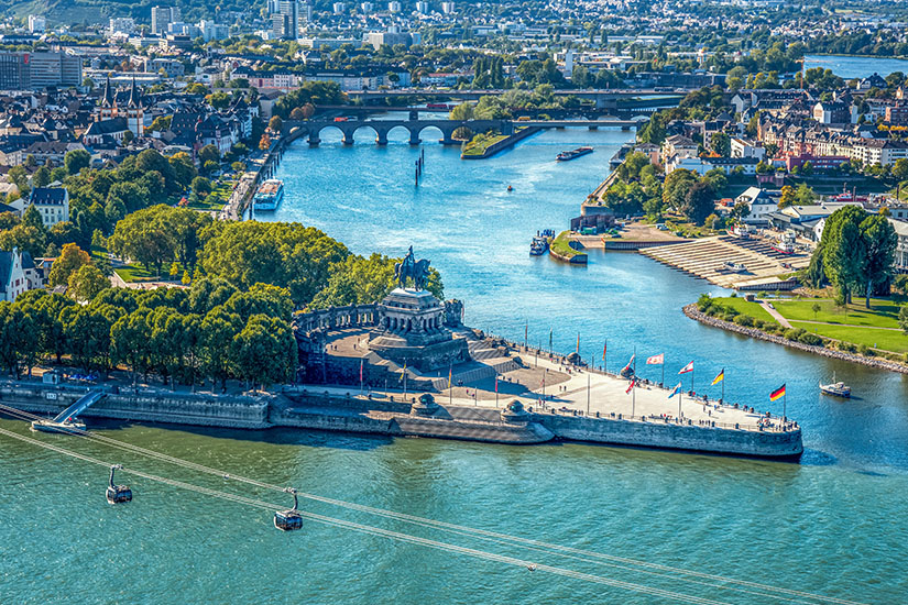 Eine Landzunge mit einem großen Reiterdenkmal ragt in den Zusammenfluss zweier Flüsse hinein. Rundherum wehen mehrere Flaggen, Spazierwege und Grünflächen säumen das Areal. Im Hintergrund sind Brücken, Schiffe und die Stadtlandschaft zu sehen. Koblenz Deutsches Eck