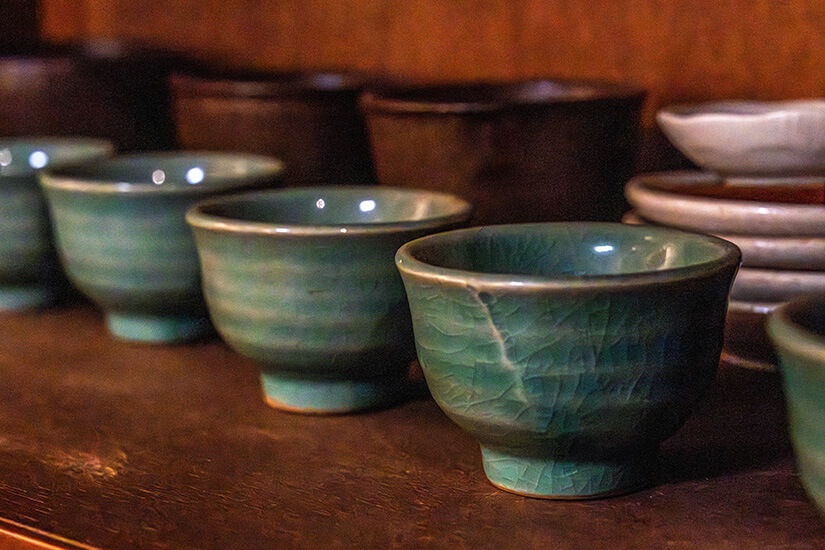 Mehrere kleine Keramikschalen in grünlich-blauer Glasur stehen in einer Reihe auf einem Regal. Im Hintergrund sind weitere Schalen und Teller gestapelt, leicht unscharf. Souvenirs Korea Keramik