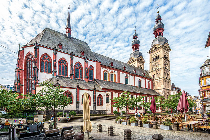 Eine große historische Kirche mit roten und weißen Fassadenelementen und zwei markanten Türmen dominiert einen offenen Platz. Davor stehen Tische, Stühle und Sonnenschirme eines Cafés, dazu einige Bäume in Pflanzkübeln. Koblenz Altstadt