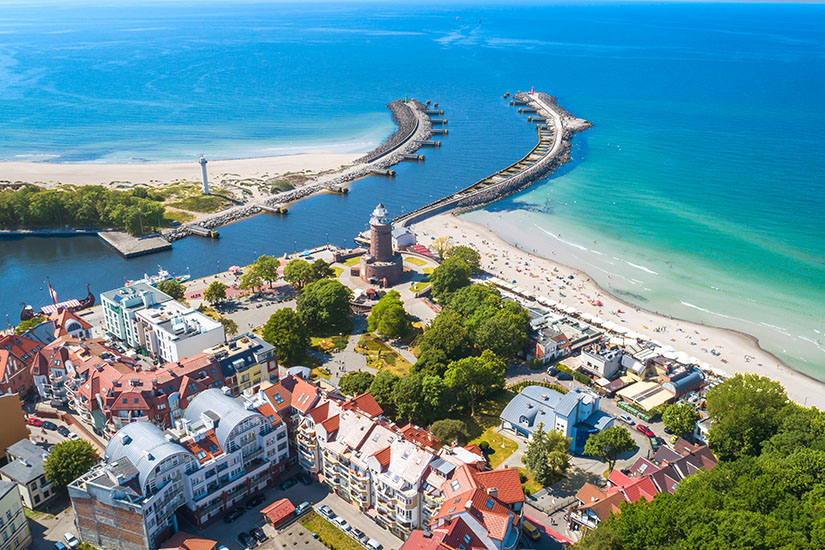 Luftaufnahme von Kolberg mit breitem Sandstrand, türkisblauem Wasser und einer Hafenmündung. Zwei lange Molen schützen den Eingang, daneben steht ein markanter Leuchtturm. Im Vordergrund sieht man die Dächer der Stadt und viel Grün entlang der Promenade.. Polen Ostsee Kolberg