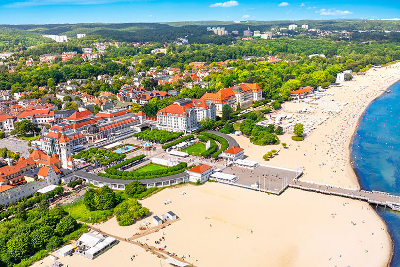 Langer, heller Sandstrand mit einer Seebrücke und einer Promenade. Dahinter liegen Gebäude mit roten Dächern, umgeben von viel Grün. Im Hintergrund ziehen sich Wälder und Wohnviertel bis zum Horizont Polen Ostsee Sopot