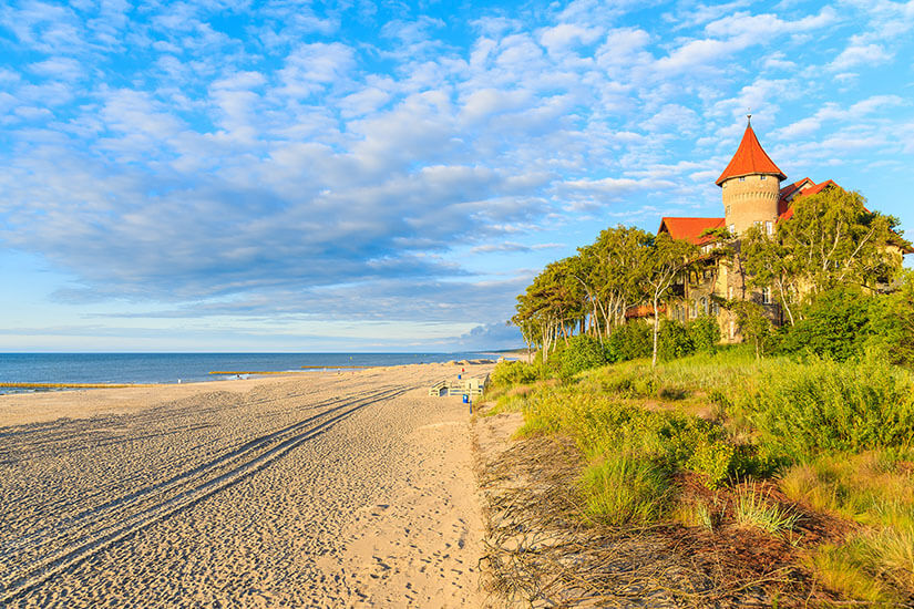 Breiter Sandstrand mit ruhigem Wasser und weitem Horizont. Dünen mit Gras, Bäume und ein historisches Gebäude mit rotem Turmdach in Leba. Polen Ostsee Leba