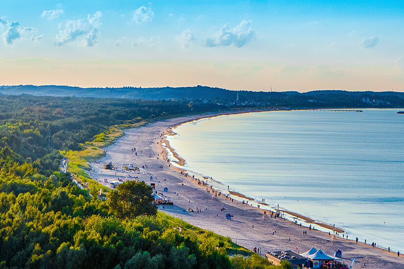 Eine lange, geschwungene Strandbucht mit ruhigem Wasser und weitem Horizont in Swinemünde. Entlang des Ufers liegen breite Sandflächen, dahinter beginnt ein grüner Küstenwald. Polen Ostsee Swinemünde