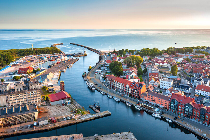 Die Hafenmündung führt direkt ins Meer, flankiert von Molen und einem Leuchtturm. Altstadt mit roten Dächern und eine schöne Uferpromenade und Boote im Hafen. Polen Ostsee Ustka