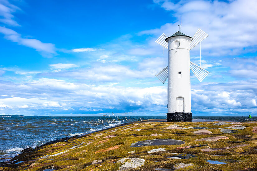 Leuchtturm mit Windradflügeln auf einer steinigen Mole direkt am Wasser. Um ihn herum schlagen Wellen gegen die Küste. Polen Ostsee Swinemünde Leuchtturm