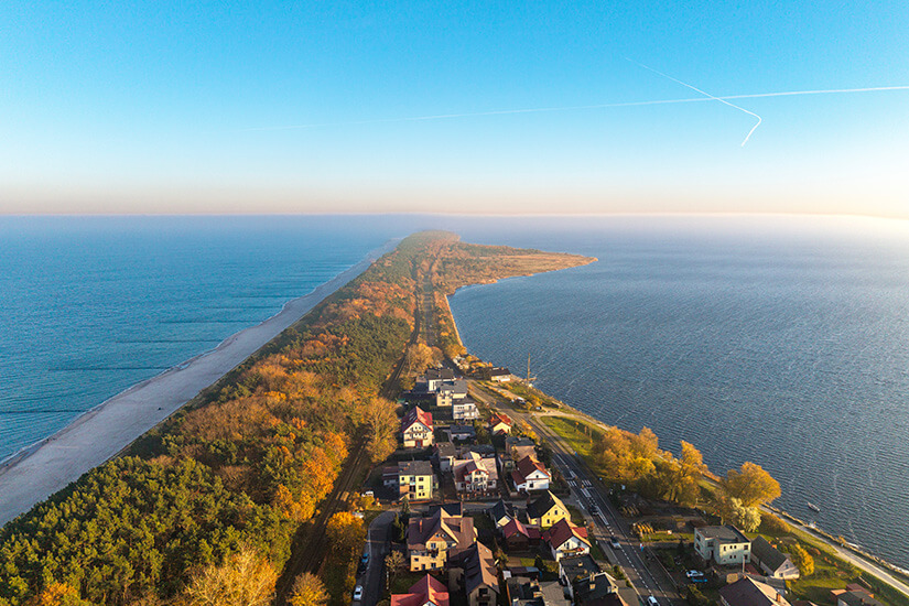 Extrem schmale Landzunge in Hel, die sich wie ein Band zwischen zwei Wasserflächen bis zum Horizont zieht. Unten liegen Häuser und eine Straße, daneben beginnt Wald und ein langer Strand. Polen Ostsee Hel