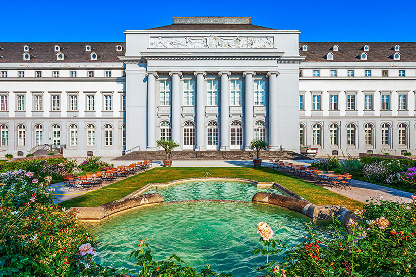 Ein großes, klassizistisches Schloss mit heller Fassade und hohen Säulen steht symmetrisch im Hintergrund. Davor liegt ein rechteckiger Brunnen mit grünlichem Wasser, umgeben von Blumenbeeten und Gartenwegen. Koblenz Kurfuerstliches Schloss