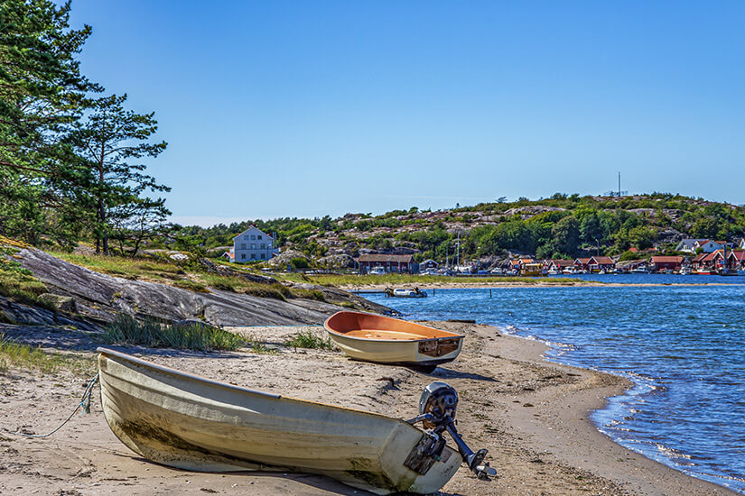 Zwei kleine Ruderboote mit Außenbordmotor liegen auf dem Sandstrand einer ruhigen Bucht, während im Hintergrund typisch schwedische Holzhäuser in Rot und Gelb zwischen glatten Granitfelsen und Kiefern hervorlugen. Schweden Schaeren Kosterinseln