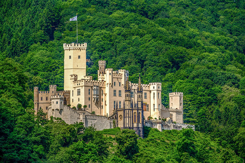 Eine märchenhafte Burg mit Türmen und Zinnen thront auf einem bewaldeten Hügel. Die hellen Mauern heben sich stark vom dichten, grünen Wald ab. Eine Fahne weht auf dem höchsten Turm. Rhein Stolzenfels