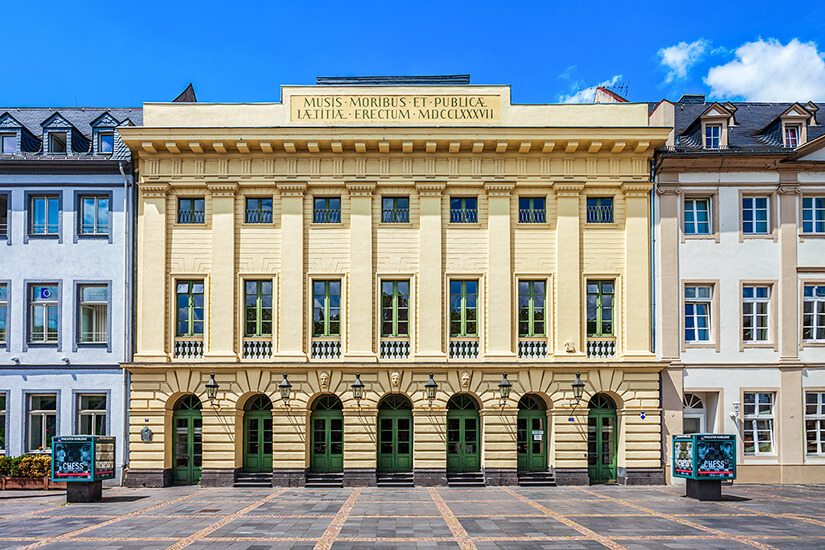 Ein klassizistisches Gebäude mit heller Fassade, Säulen und symmetrischer Fensteranordnung steht an einem gepflasterten Platz. Über dem Dachgesims ist eine lateinische Inschrift angebracht. Seitlich sind Plakatsäulen und benachbarte Häuser mit ähnlicher Höhe zu sehen. Koblenz Theater