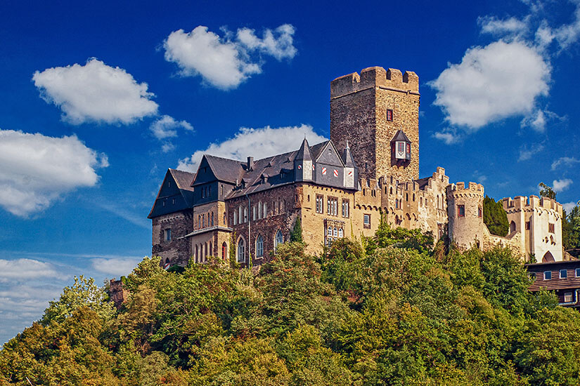 Eine mittelalterliche Burg thront auf einem bewaldeten Hügel mit Zinnen, Türmen und einem markanten Bergfried. Die steinernen Fassaden heben sich klar vom satten Grün der Bäume ab. Darüber spannt sich ein tiefblauer Himmel mit vereinzelten weißen Wolken. Rheintal Lahneck
