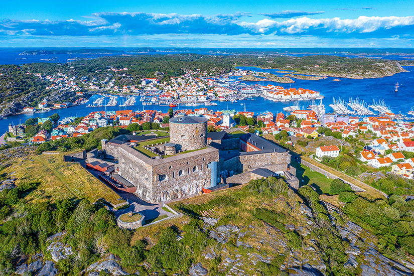Eine mächtige Steinfestung thront auf einem Felsen über einer malerischen Küstenstadt mit weißen Häusern, orangefarbenen Dächern und einem geschäftigen Yachthafen, umgeben von der typischen schwedischen Schärenlandschaft. Schweden Schaeren Marstrand