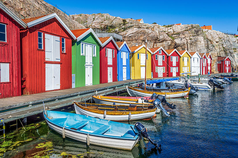 Knallbunte Hütten in Rot, Grün, Blau und Gelb reihen sich entlang eines Holzstegs, vor dem kleine Motorboote im klaren Wasser dümpeln, während glatte Granitfelsen den Hintergrund bilden. Schweden Schaeren Smoegen