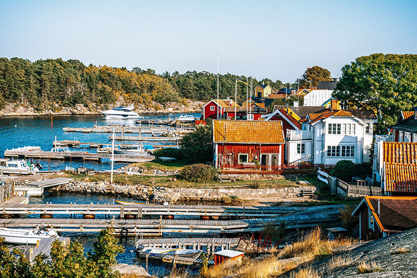 Charmantes Schärendorf mit bunten Holzhäusern in traditionellem Rot und Orange, verwitterten Holzstegen und kleinen Booten, die im ruhigen Wasser vor einer bewaldeten Felsenkulisse liegen. Schweden Schaeren Sandhamn