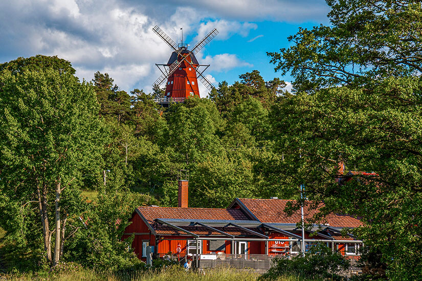 Während im Vordergrund ein typisch schwedisches Holzhaus im charakteristischen Falunrot zwischen den Bäumen hervorlugt, ragt eine traditionelle rote Windmühle über den Baumwipfeln eines üppig grünen Waldes empor. Schweden Schaeren Utoe