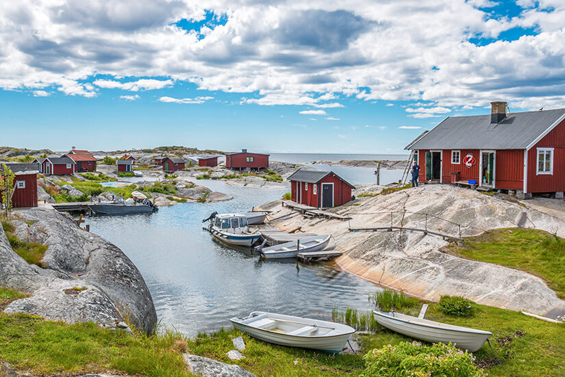 Verstreute rote Holzhütten ducken sich zwischen glatten Granitfelsen auf einer kargen Schäreninsel, während kleine Boote in einem natürlichen Felsenkanal vertäut liegen und das offene Meer am Horizont schimmert. Huvudskaer Schweden