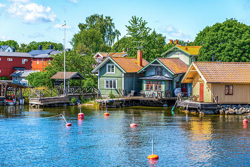 Pastellfarbene Holzhäuser in Grün, Gelb und traditionellem Rot reihen sich malerisch am Wasser entlang, mit eigenen Stegen und Bootshäusern, während orangefarbene Bojen im ruhigen Wasser schaukeln. Schweden Schaeren Vaxholm