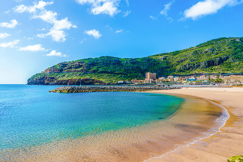 Der Praia de Machico ist ein wunderschöner Sandsrtand direkt an der Promenade. Direkt hinter der Bucht sind Häuser und Berge sichtbar. Machico Portugal Strand