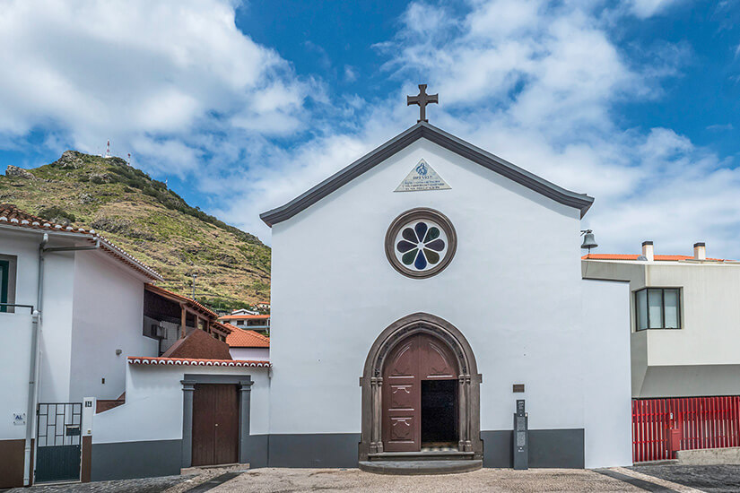 Die weiße Igreja Matriz de Machico mit ihrem Turm und der schlichten Fassade steht im Zentrum von Machico. Davor liegt ein gepflasterter Platz mit Bänken, Bäumen und niedrigen Mauern. Im Hintergrund sind weitere Gebäude der Innenstadt zu sehen. Machico Portugal Igreja