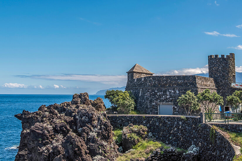 Die Festung Forte São João Baptista an der Küste von Porto Moniz auf Madeira. Der steinerne Bau sitzt auf einem dunklen, felsigen Untergrund direkt über dem Atlantik, mit massiven Mauern und klar erkennbaren Kanten. Im Hintergrund liegen Meer und Horizont, daneben die Küstenlinie. Machico Portugal Forte São João Baptista