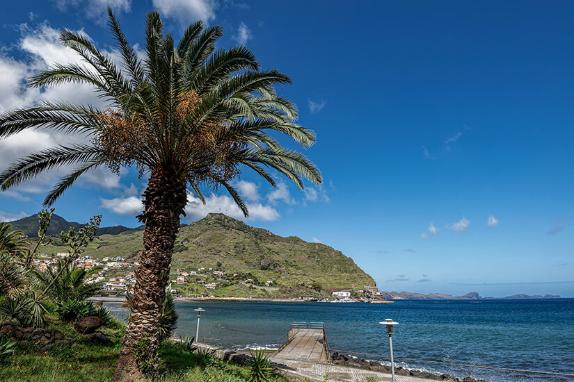 Die Stadt zieht sich vom Ufer entlang des Tals nach hinten, eingerahmt von grünen Hängen. Promenade, Strandbereich und die Gebäude im Zentrum liegen nah beieinander, dahinter sind die Berge. Machico Portugal Meer