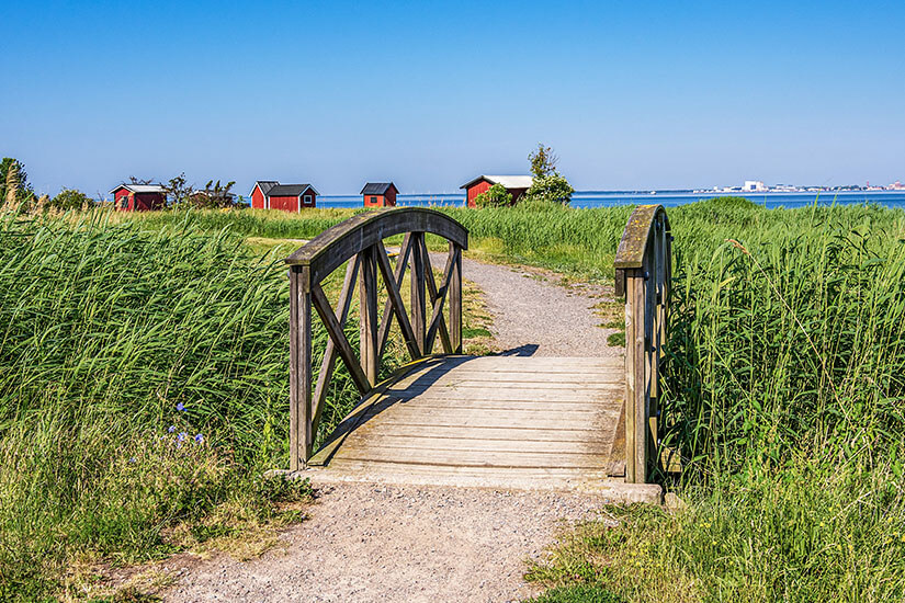 Eine kleine Holzbrücke mit dekorativem Geländer führt über einen Graben durch hohes Schilf, während der Kiesweg zu verstreuten roten Fischerhütten am Meer hinführt. Schweden Schaeren Oeland