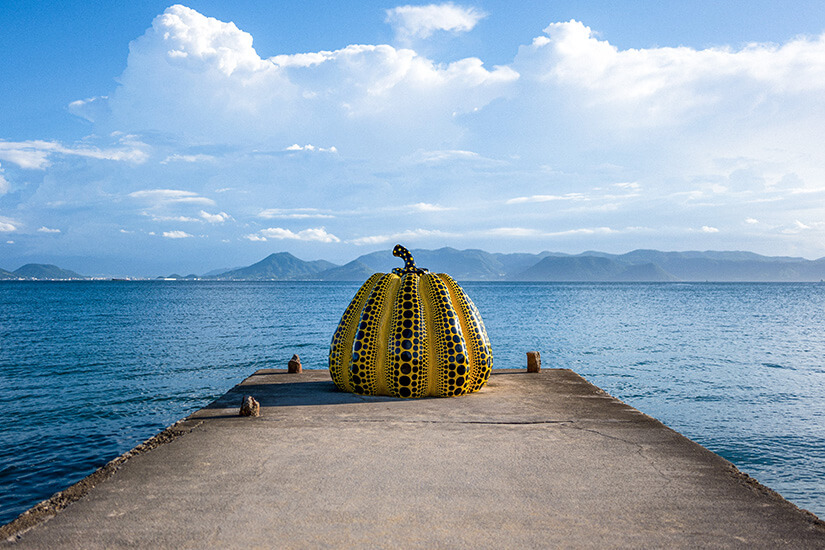 Eine gelbe Kürbis-Skulptur mit Punkten sitzt am Ende eines Piers am Meer. Im Hintergrund ist die Kulisse einer Insel zu sehen. Naoshima Japan Kuerbis Skulptur