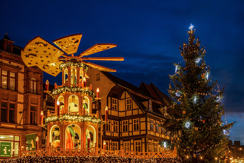 Ein festlich beleuchteter Weihnachtsmarkt mit einer großen, mehrstöckigen Weihnachtspyramide voller Figuren und Kerzen. Daneben steht ein geschmückter Tannenbaum mit Lichtern vor historischen Fachwerkhäusern. Quedlinburg Weihnachtsmarkt