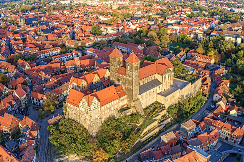Eine historische Altstadt mit dicht gedrängten Fachwerkhäusern und roten Ziegeldächern breitet sich rund um einen markanten Burgberg aus. Auf dem Hügel thront ein großes Kirchen- und Schlossensemble mit massiven Steinmauern, das deutlich über die Stadt hinausragt. Quedlinburg Stadt