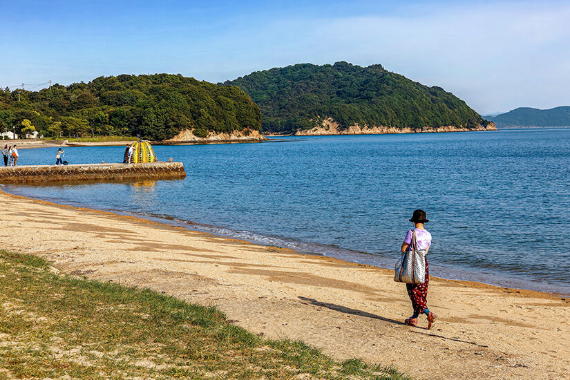Eine Frau geht am Sandstrand auf der Insel Naoshima spazieren. Das blaue Meer und die Kürbis-Skulptur am Pier sind zu sehen. Naoshima Japan Wetter