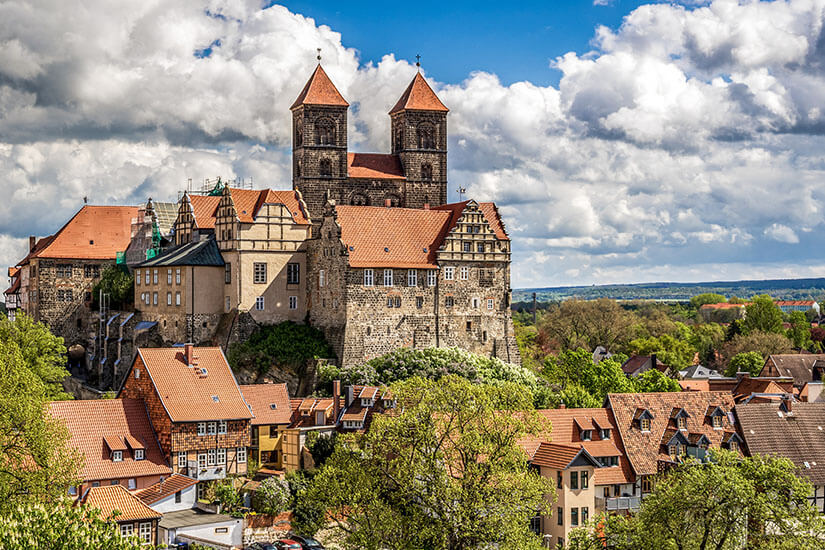 Eine große romanische Kirche mit zwei Türmen thront über einer historischen Altstadt. Darunter liegen dicht bebaute Häuser mit roten Ziegeldächern, umgeben von viel Grün. Quedlinburg Stiftskirche