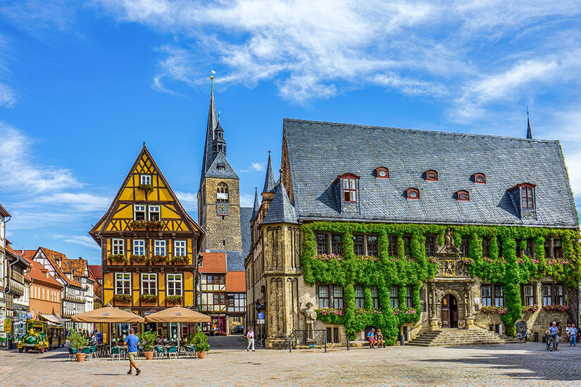 Ein historischer Marktplatz mit Fachwerkhäusern und einem großen, mit Efeu bewachsenen Rathaus. Im Hintergrund ragt ein Kirchturm über die Dächer, während Menschen über den Platz schlendern oder in Straßencafés sitzen. Quedlinburg Marktplatz