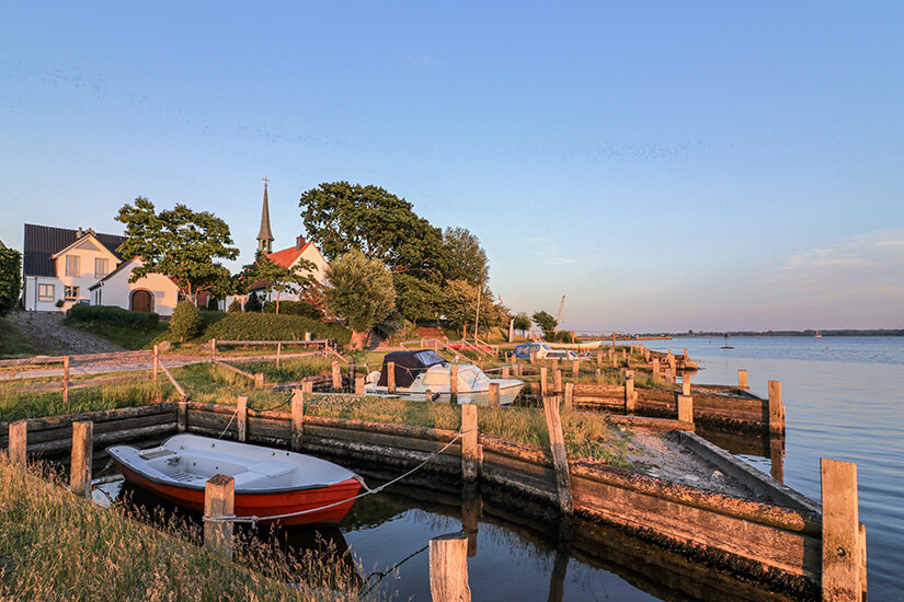 Kleines rotes Ruderboot liegt an einem hölzernen Anlegesteg im ruhigen Wasser von Maasholm vor einer weißen Kirche mit spitzem Turm bei Abendsonne. Ostsee Maasholm Geheimtipps Ostsee Maasholm