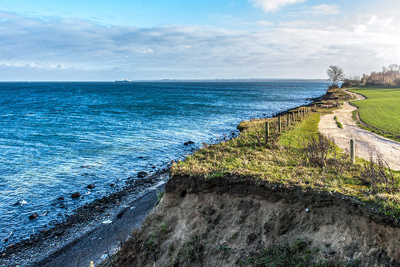 Blick entlang der Abbruchkante des Brodtener Steilufers auf die blaue Ostsee, mit einem schmalen Wanderweg oberhalb der Klippen und Feldern im Hintergrund. Geheimtipps Ostsee Brodtener Steilufer