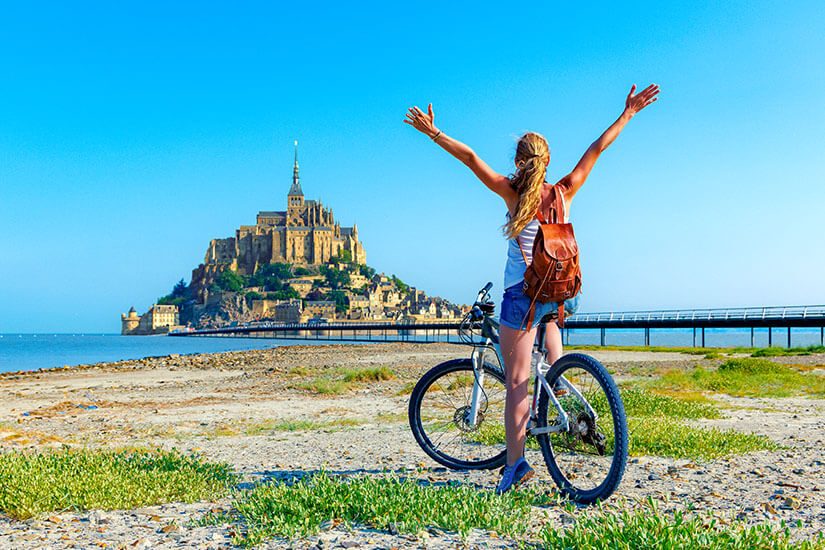 Eine Frau steht mit ausgebreiteten Armen neben ihrem Fahrrad am Strand und blickt auf eine beeindruckende Klosterinsel im Meer. Packliste Frankreich Fahrrad