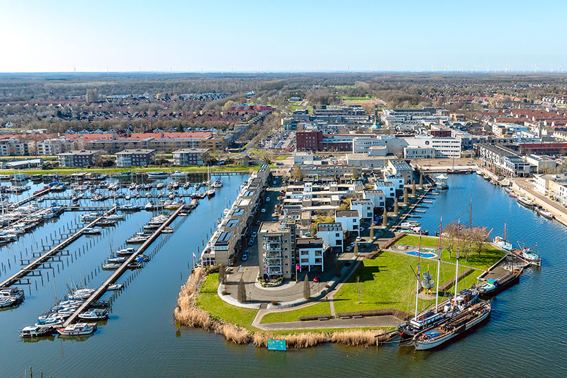Eine moderne Hafenanlage mit zahlreichen Segelbooten und Stegen erstreckt sich entlang eines Kanals. Auf einer Landzunge stehen Wohngebäude, Grünflächen und ein historisches Segelschiff am Kai. Im Hintergrund breitet sich eine flache Stadtlandschaft mit Straßen, Häusern und viel Weite bis zum Horizont aus. Flevoland Provinz