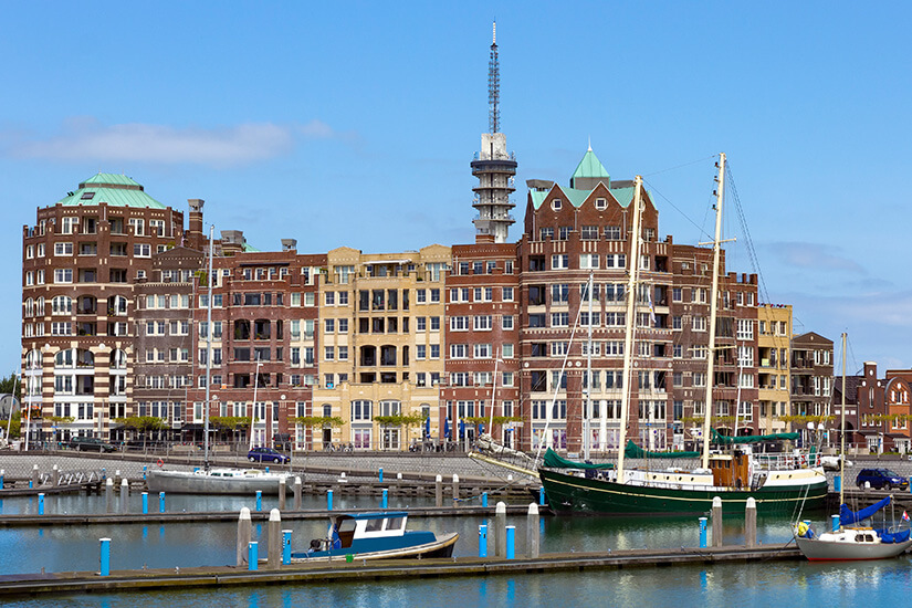 Ein Yachthafen mit mehreren Segelbooten liegt ruhig vor einer Reihe markanter Backstein- und Wohngebäude. Die Architektur wirkt modern mit maritimen Elementen und unterschiedlich hohen Fassaden. Im Hintergrund ragt ein schlanker Turm mit Antenne über die Dächer hinaus. Flevoland Lelystad