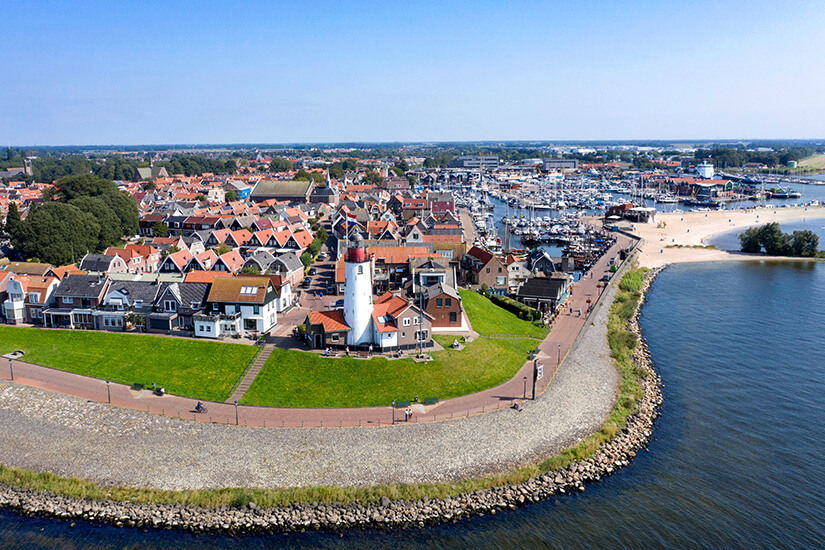Ein Küstenort mit dicht stehenden Häusern und roten Ziegeldächern erstreckt sich entlang eines Deichs. In der Mitte steht ein weißer Leuchtturm auf einer grünen Wiese nahe der Uferpromenade. Dahinter liegt ein großer Hafen mit zahlreichen Booten und Yachten. Flevoland Urk