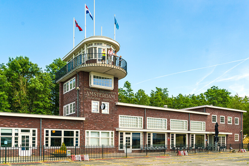 Ein Backsteingebäude mit einem runden Kontrollturm trägt die Aufschrift „Gemeente Luchthaven Amsterdam Municipal Airport“. Auf dem Balkon des Turms stehen mehrere Personen, darüber wehen Fahnen. Holland Luftfahrtmuseum