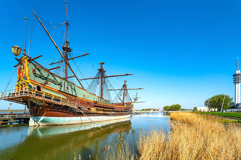 Ein historisches Segelschiff mit hohen Masten und reich verziertem Heck liegt in einem ruhigen Hafenbecken. Im Hintergrund ragt ein moderner Aussichtsturm neben grünen Uferflächen in den blauen Himmel. Holland Batavialand