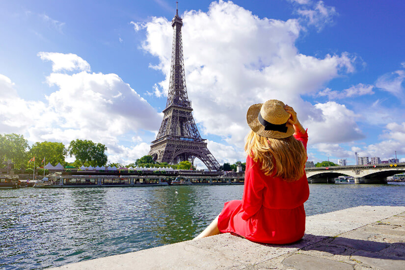 Eine Frau in rotem Kleid sitzt am Ufer der Seine und blickt auf den Eiffelturm. Der Himmel ist blau mit großen weißen Wolken, die die Szene hell und freundlich wirken lassen. Packliste Frankreich Paris