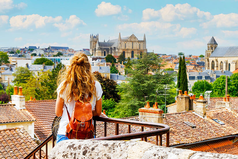 Eine Frau mit Rucksack steht auf einer Aussichtsterrasse und blickt über die Dächer einer historischen Stadt. Im Hintergrund erhebt sich eine große Kathedrale über den roten Ziegeldächern. Packliste Frankreich Ausblick