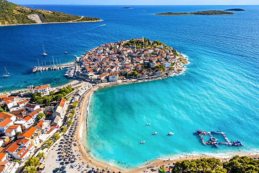 Kleine Altstadt auf einer Landzunge mit roten Ziegeldächern in Kotor, die von zwei Buchten umgeben ist. Vorne schimmert das Wasser türkis und flach, am Rand liegen Sonnenschirme am gebogenen Strand und ein kleiner Yachthafen mit Booten. Dahinter wird das Meer dunkler und weit, mit Inseln am Horizont und grünen Hügeln links und rechts. Strände Montenegro Kotor